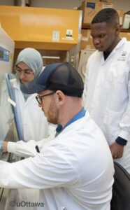 Trois personnes travaillent ensemble dans un laboratoire. La personne au premier plan, portant une casquette et des lunettes de sécurité, manipule du matériel derrière une hotte de sécurité biologique. À gauche, une collègue portant un hijab bleu clair et des lunettes observe attentivement l’expérience. À droite, une troisième personne en sarrau blanc se tient debout et regarde la manipulation avec sérieux. L’arrière-plan montre un environnement de laboratoire avec des boîtes et équipements. © uOttawa
